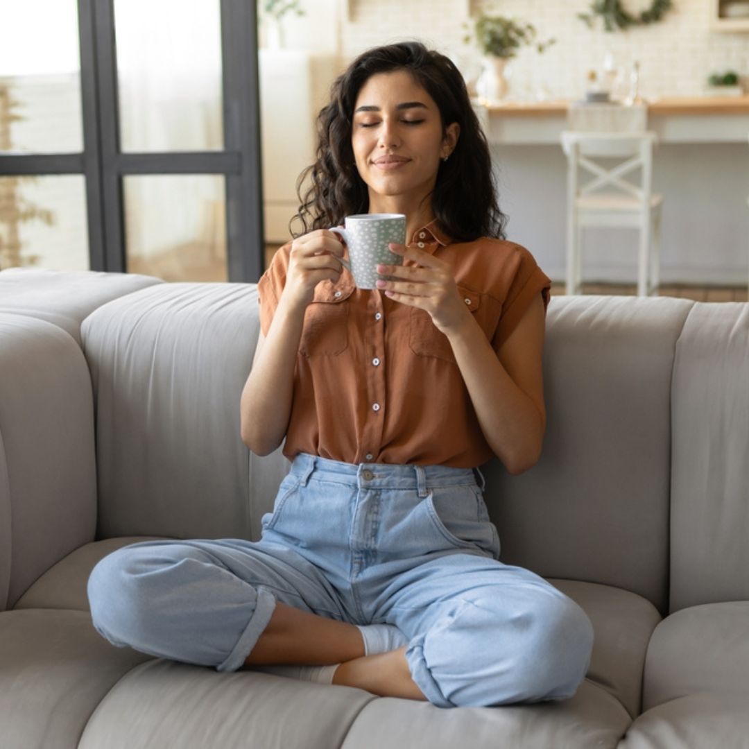 Eine junge Frau sitzt auf einer Couch und trinkt eine Tasse Kaffee.
