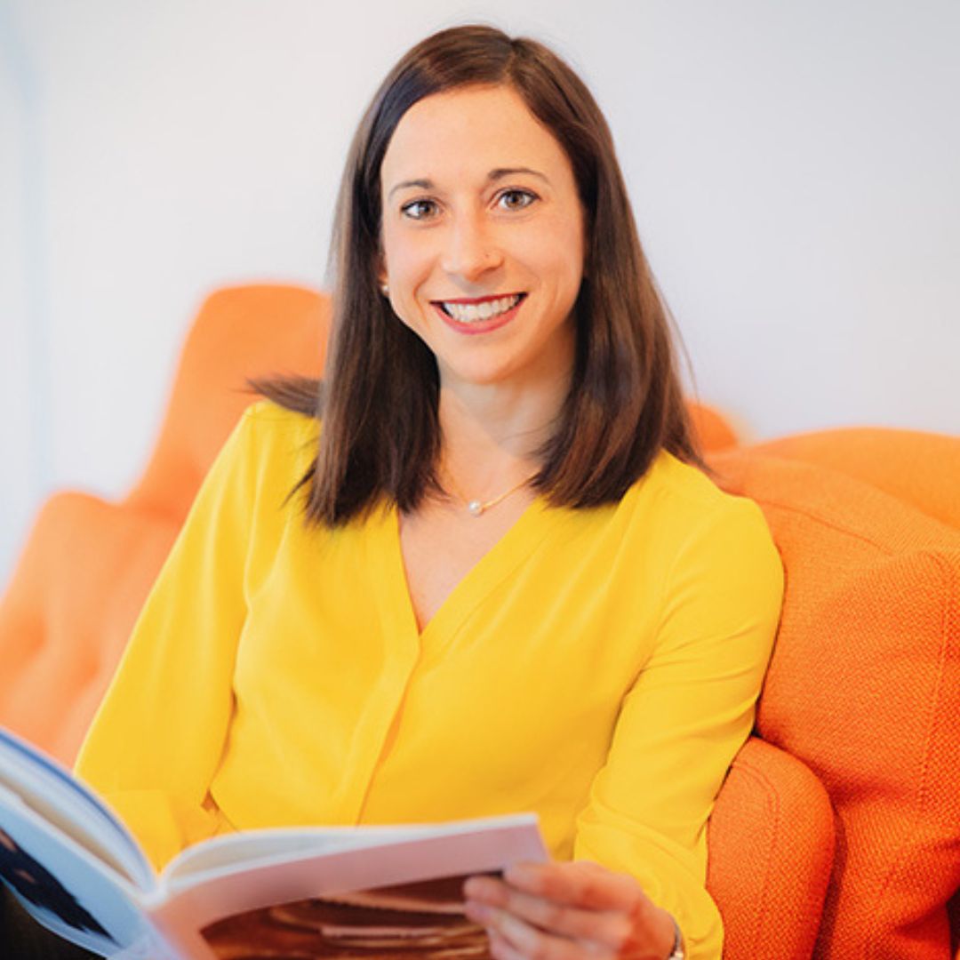 Eine Frau sitzt auf einer orangefarbenen Couch und liest eine Zeitschrift.